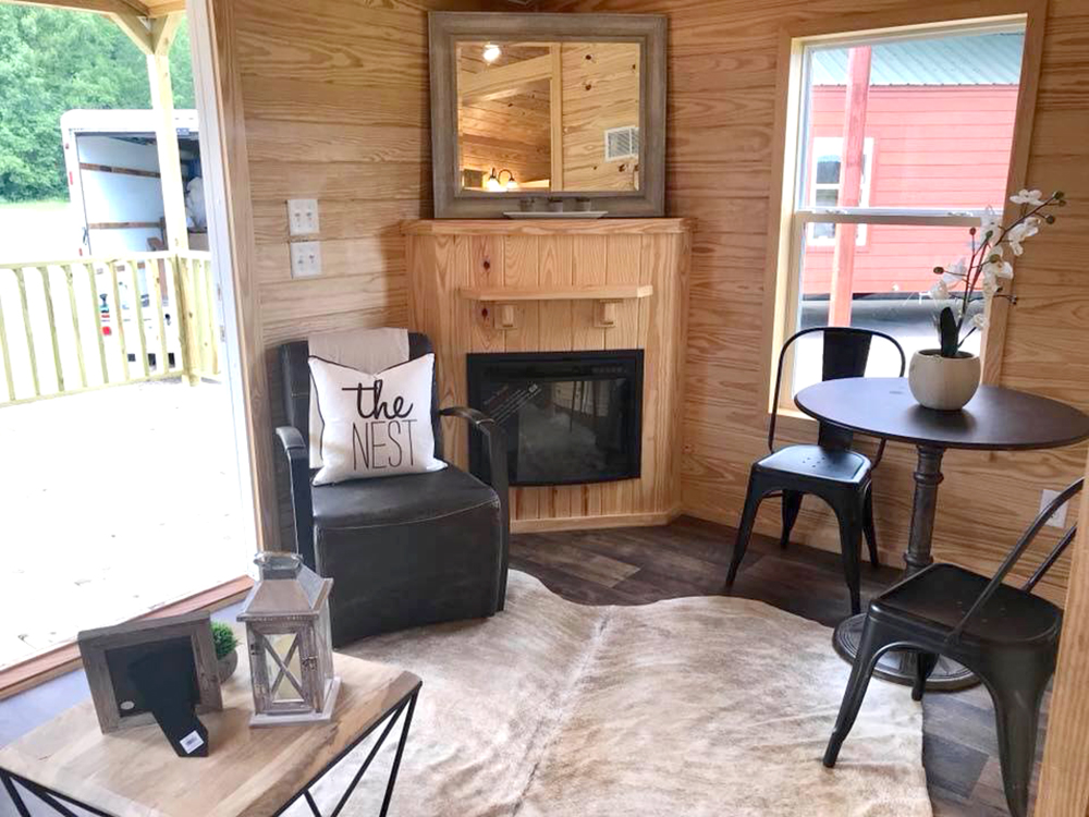 Cozy wooden room with a corner fireplace, black armchair, and cushion labeled "The Nest." Round table with two chairs and a white orchid adds warmth.