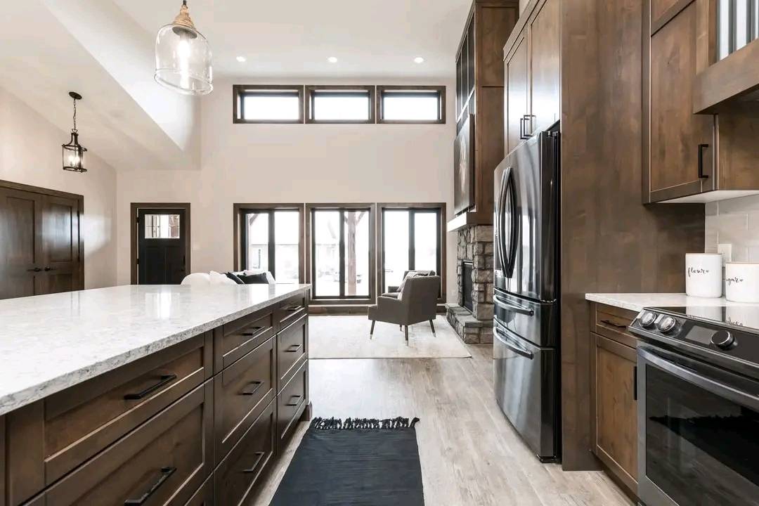 Modern kitchen with earthy tones, featuring dark wood cabinetry, stainless steel appliances, and a white countertop. Large windows and high ceiling add brightness.