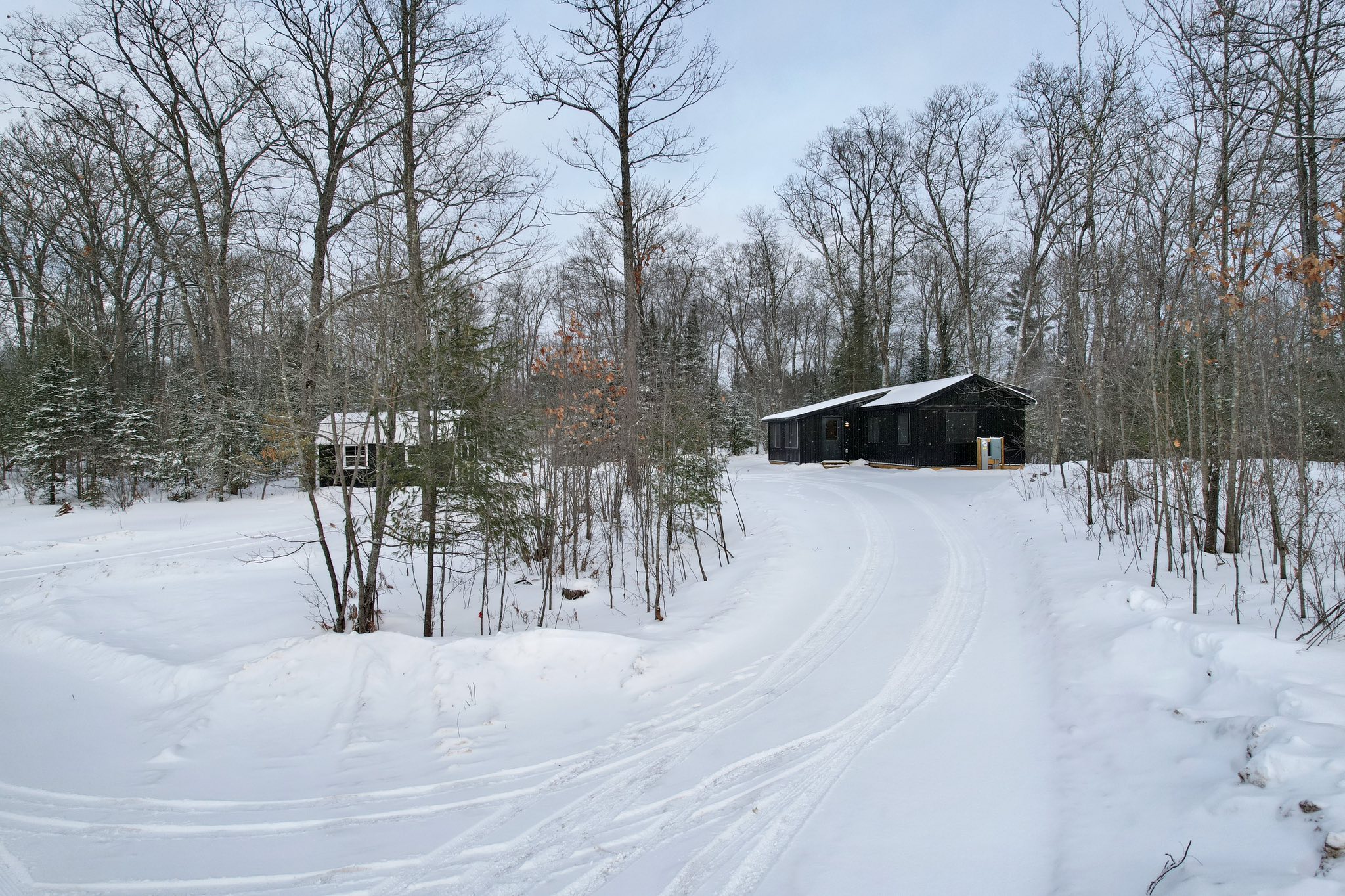Snowy forest scene with a dark cabin and a smaller structure surrounded by bare trees. The snow-covered path creates a serene, winter ambiance.