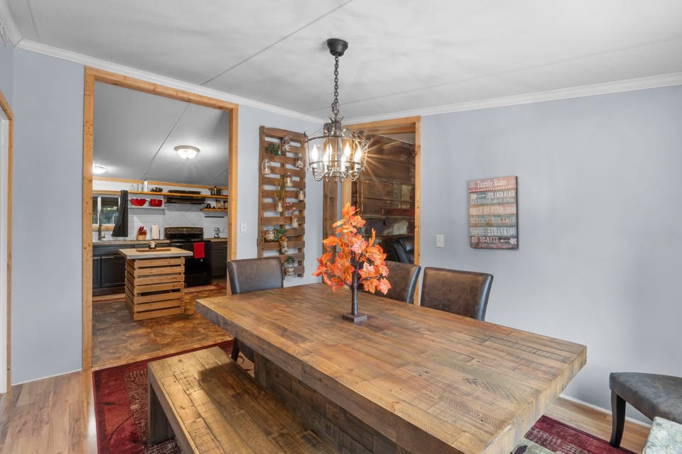 A cozy dining room features a rustic wooden table with a fall-themed centerpiece. A warm light fixture hangs above, leading to a kitchen with wood accents.