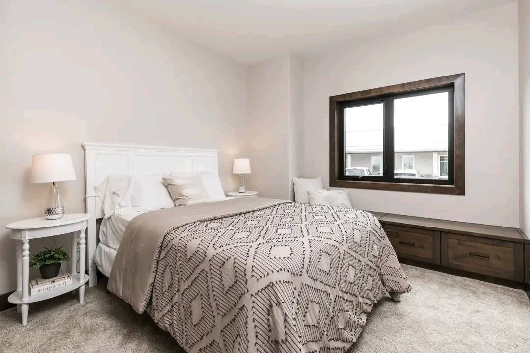 Cozy bedroom with a large bed featuring a patterned beige comforter, flanked by two white nightstands with lamps. Soft light, neutral walls, and a window seat add warmth.