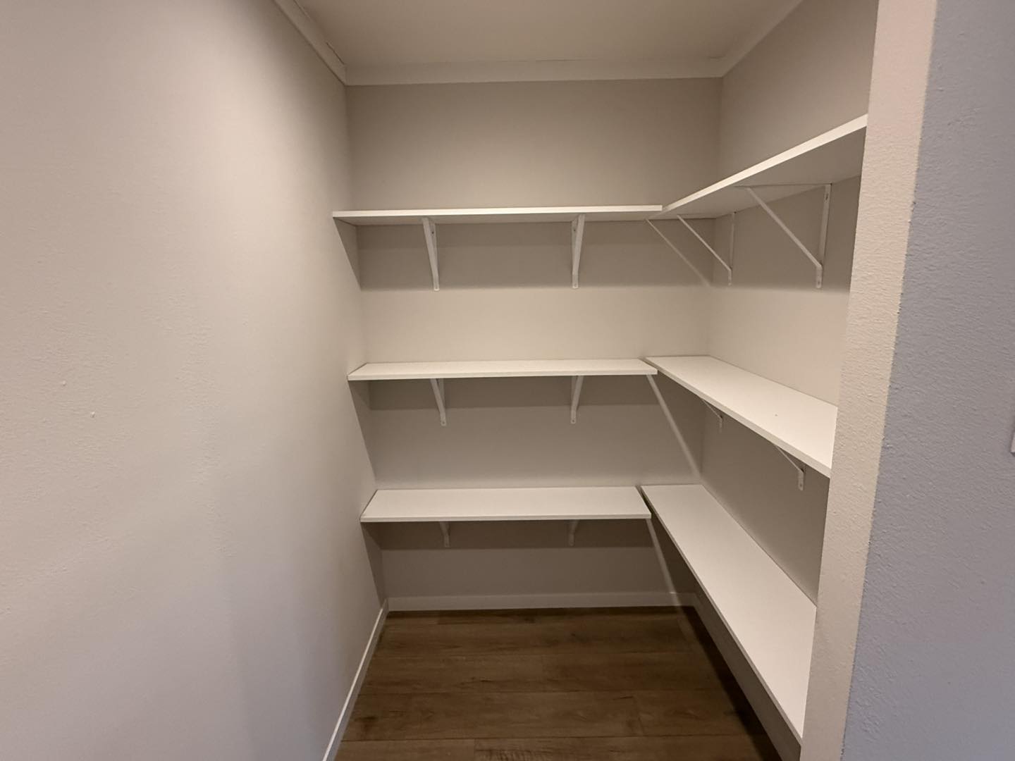 Empty pantry with white L-shaped shelves on the left and right walls. The walls are a soft beige color, and the floor is light wood, creating a clean and organized appearance.