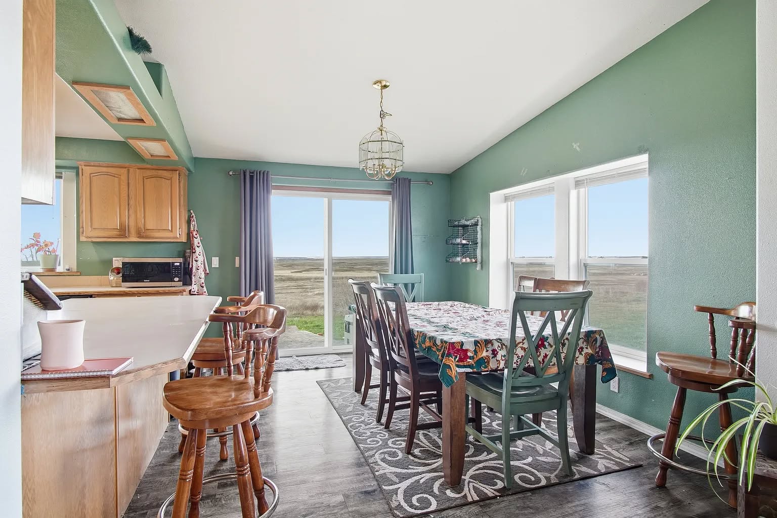 Dining room with green walls, wooden table, and eight chairs. Bright window view of open fields. Cozy atmosphere with a patterned rug and hanging light fixture.