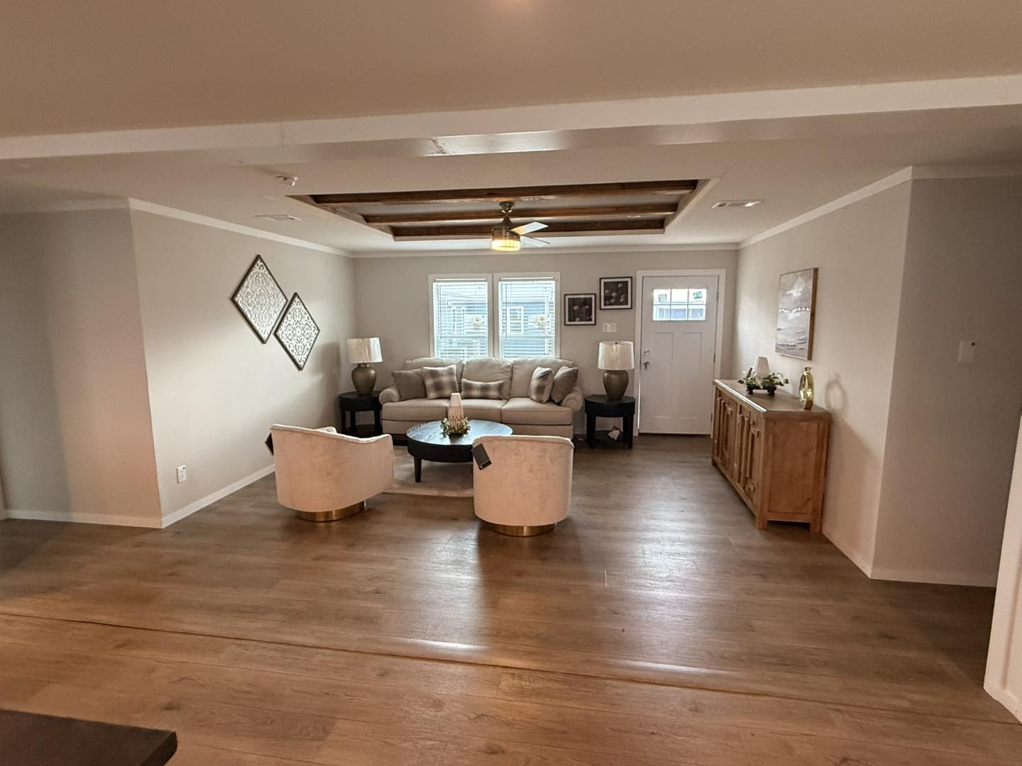 Cozy living room with beige tones, featuring a plush sofa, two armchairs, and wooden furniture. Light from a ceiling fan accents the warm, inviting space.