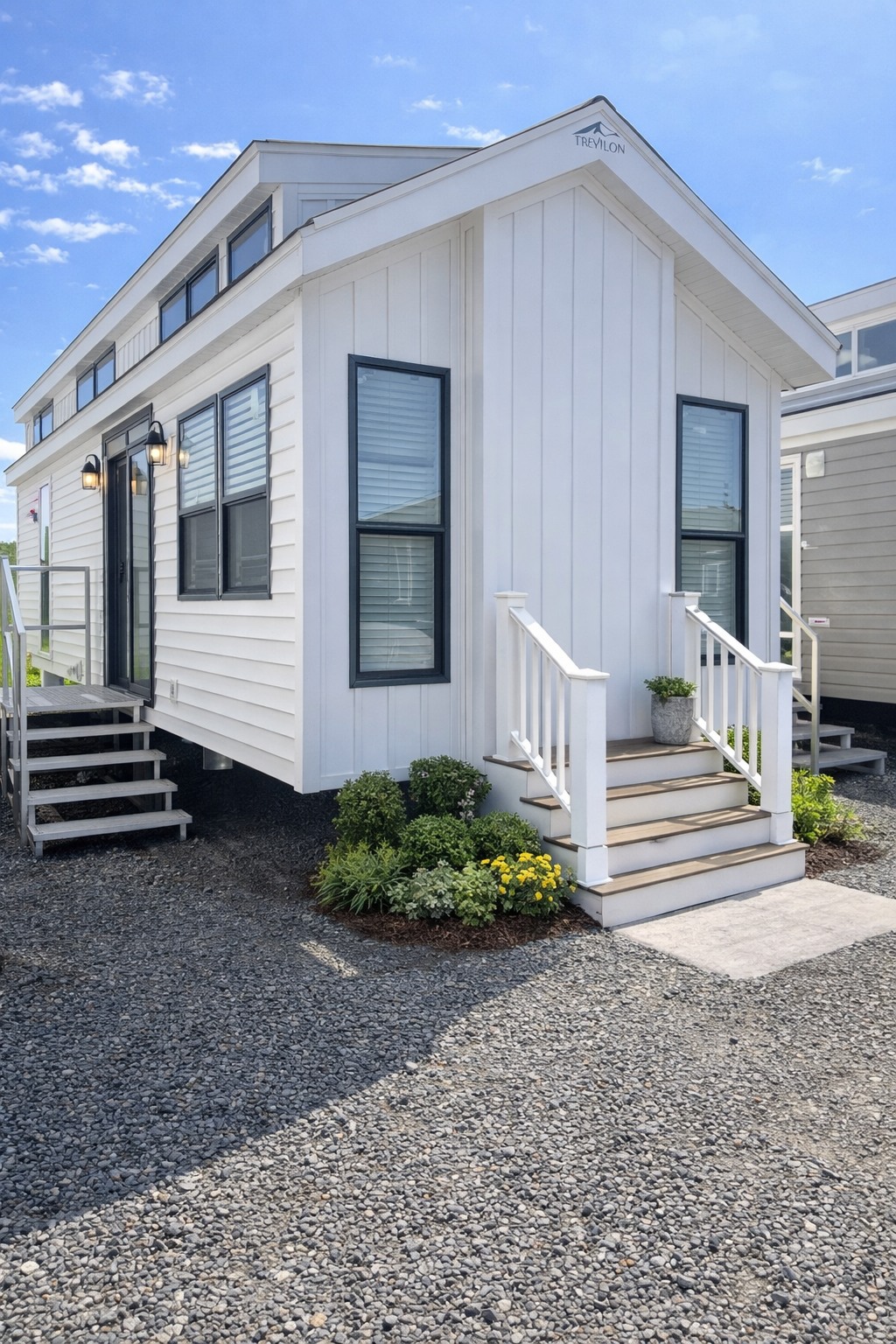 White tiny house with black-trimmed windows, two entry steps, and potted plants. Gravel path and clear blue sky create a serene, modern setting.