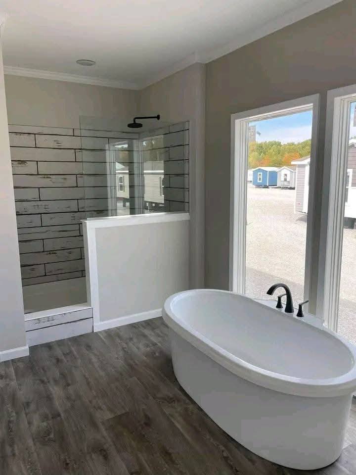 A modern bathroom with a freestanding oval bathtub near large windows. To the left, a walk-in shower features rustic wooden tiles. The tone is serene and minimalistic.