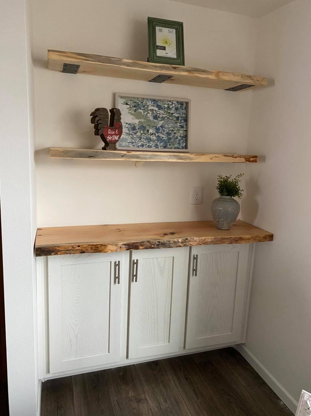 A cozy nook features white cabinets with a raw wood countertop. Above, two wooden shelves display framed art, a potted plant, and decor.