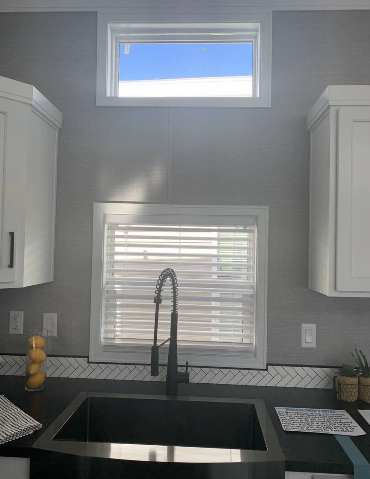 Modern kitchen sink area with black faucet and countertop. Two windows with blinds provide natural light. White cabinets contrast the dark counters.