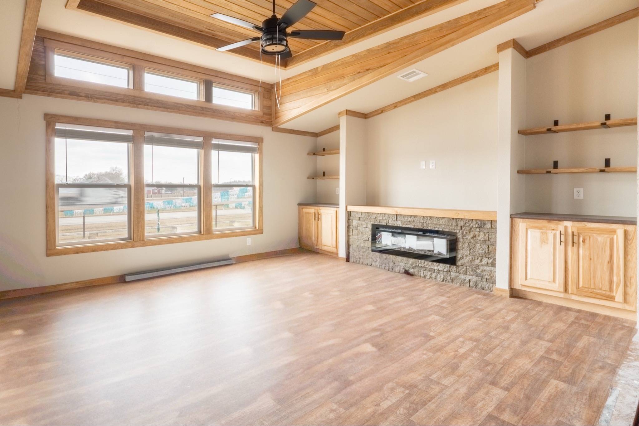 Spacious living room with light wood floors, large windows, modern electric fireplace, wooden ceiling accents, open shelves, and natural tones.