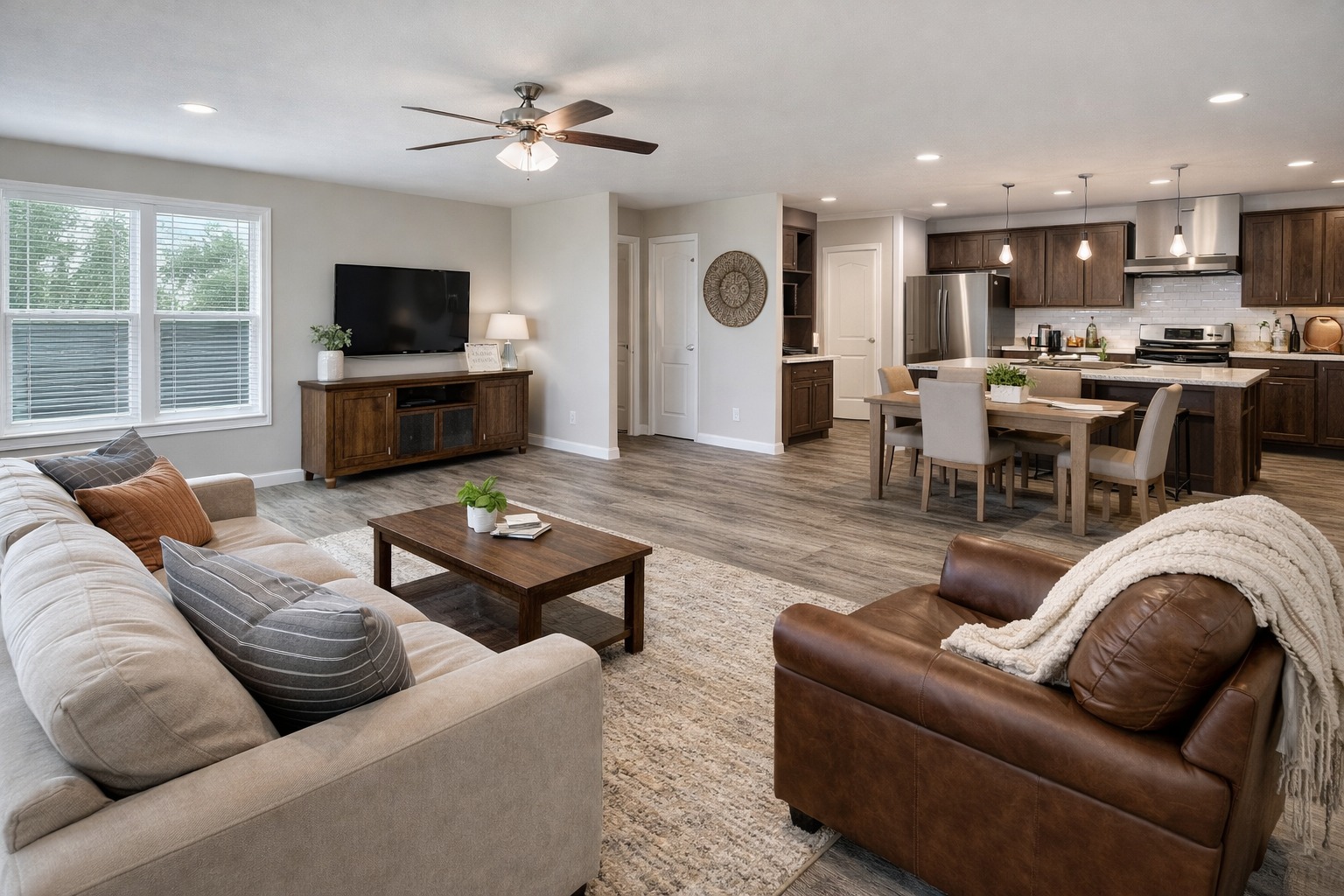 Open-concept living room and kitchen with neutral tones. Cozy sofas and armchair, wooden coffee table, TV, dining area, pendant lights, and modern appliances.