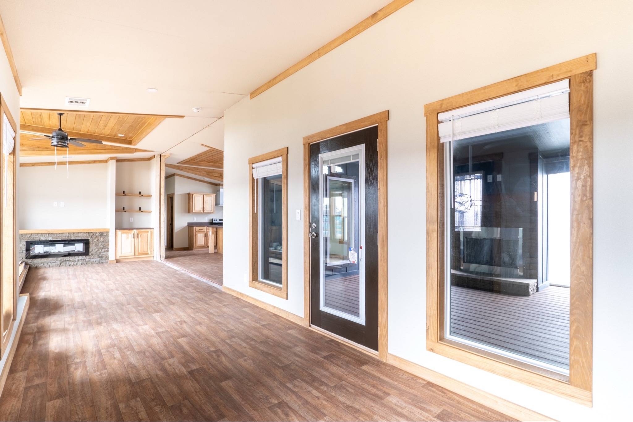 Spacious interior with wooden flooring and trim, large windows, and a glass door. A ceiling fan and fireplace enhance the cozy yet modern feel.