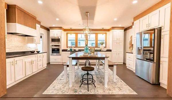 Spacious kitchen with white cabinets, a central wooden table, and stainless steel appliances. A large rug and bright windows create a welcoming vibe.