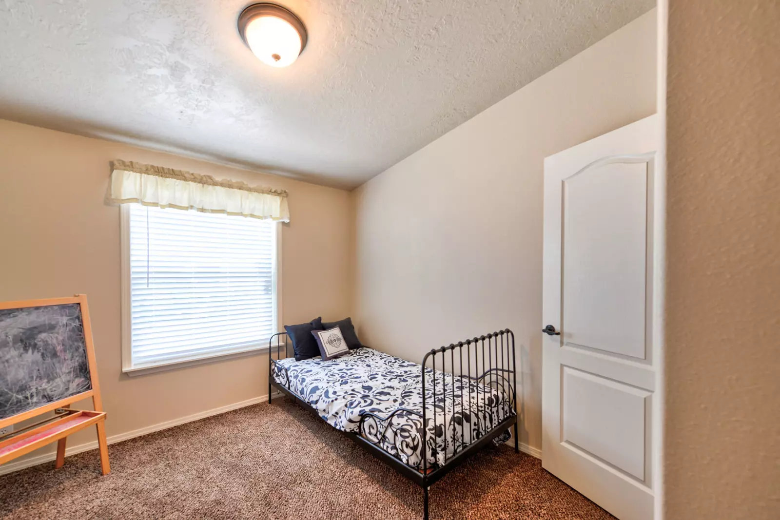 A simple bedroom with beige walls and a single ornate metal bed with patterned bedding. A window with white blinds and a chalkboard stand nearby. Cozy and minimalistic.