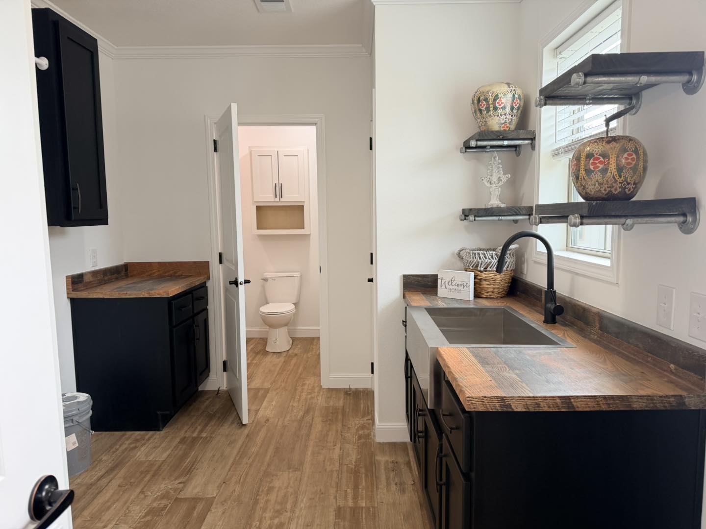 Laundry room with rustic wood countertops, black cabinets, and industrial shelves holding decorative vases. A sink is under a window. A door to a small bathroom with a visible toilet and cabinet is open. The setting is clean and modern.