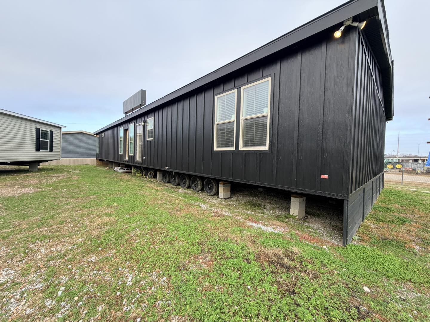 A black, rectangular mobile home on wheels sits on a grassy lot. The sky is overcast. Windows line the side, giving a modern and functional look.