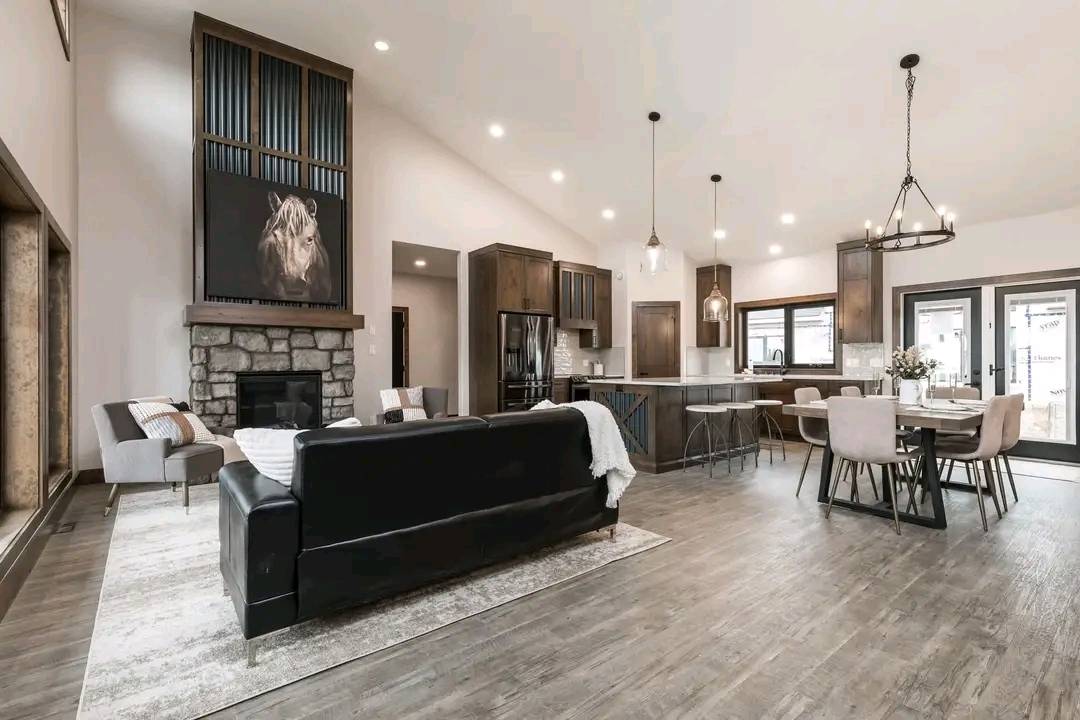 Modern open-concept living room and kitchen with high ceilings. Features a stone fireplace, dark leather sofa, dining table, and wood accents, creating a cozy, elegant ambiance.