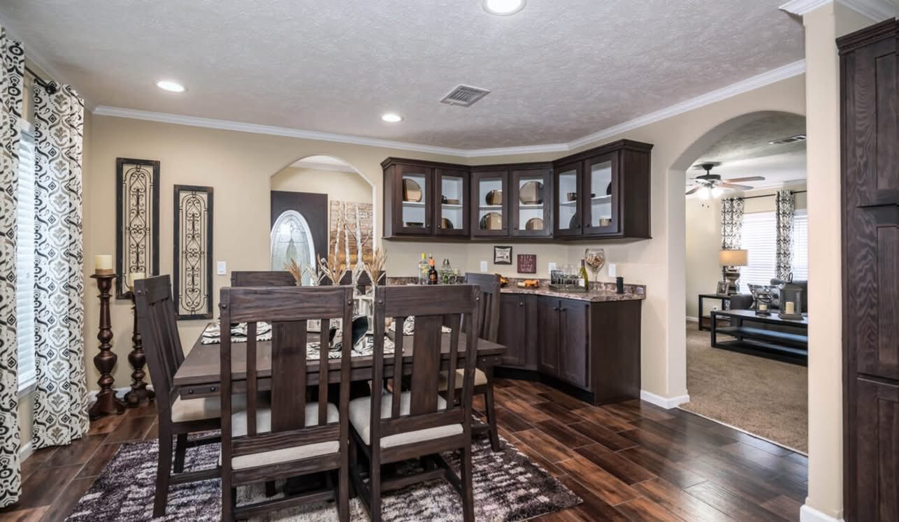 A cozy dining room with a wooden table set for six, surrounded by elegant decor and dark cabinets. An archway leads to a warmly lit living room.