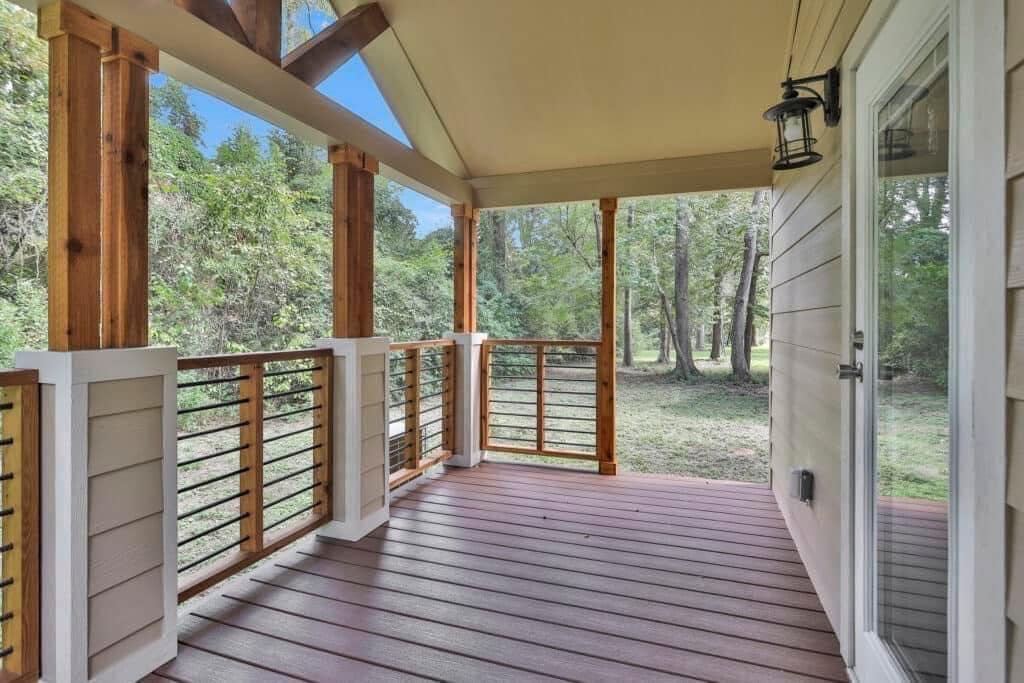 A serene porch with wooden railings and a sloped roof overlooks a lush, green wooded area. A glass door and rustic lantern enhance the cozy, natural atmosphere.