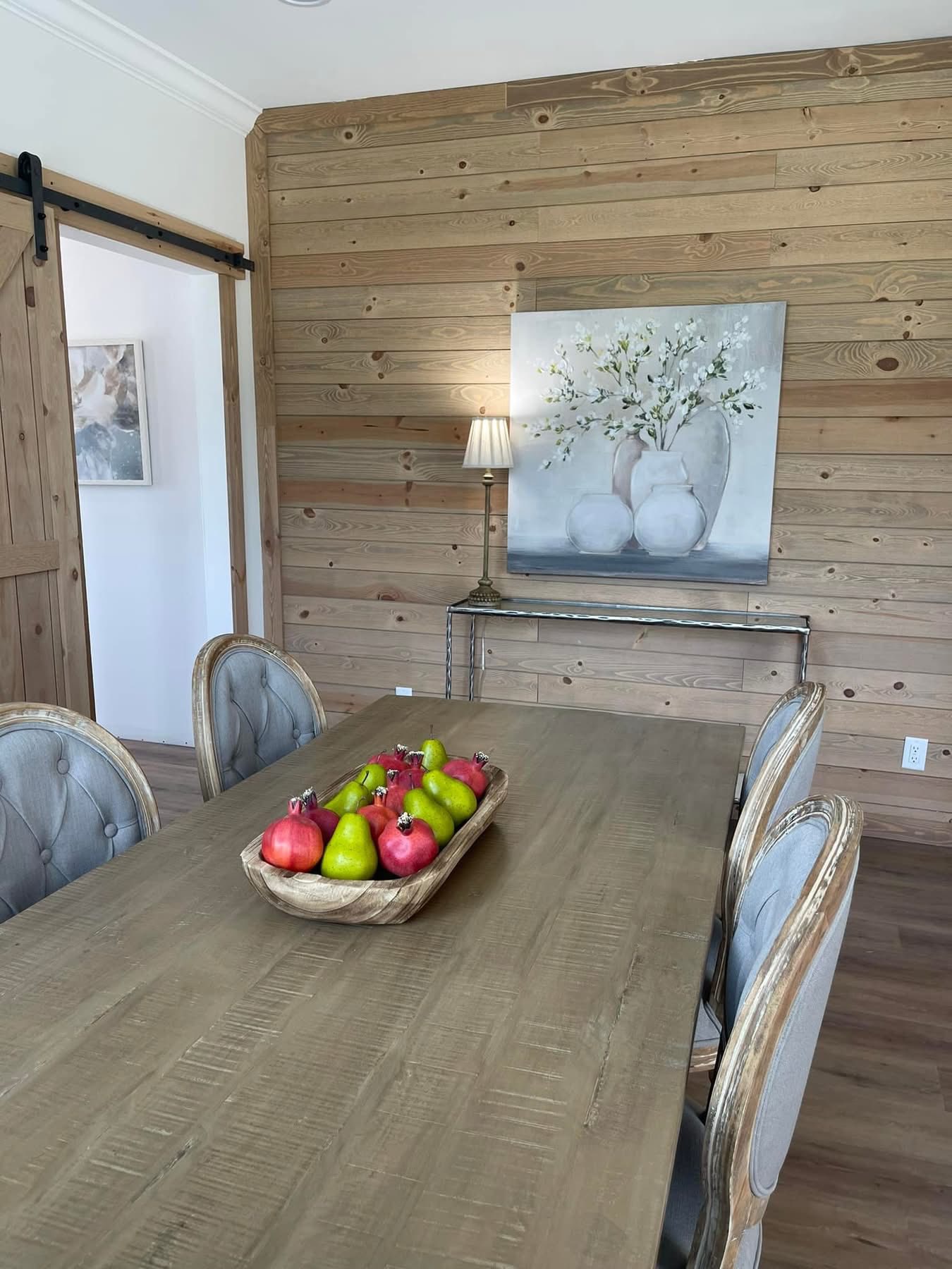 Dining room with a rustic wooden table and six chairs. A centerpiece of red apples and green pears decorates the table. A painting and lamp on a side table along a wood-paneled wall create a cozy atmosphere.