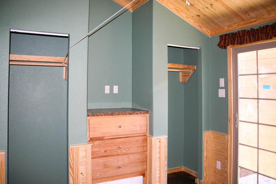 A small room with green walls, two open closets, wood paneling, and built-in drawers. A sliding glass door with shades adds a cozy, rustic feel.