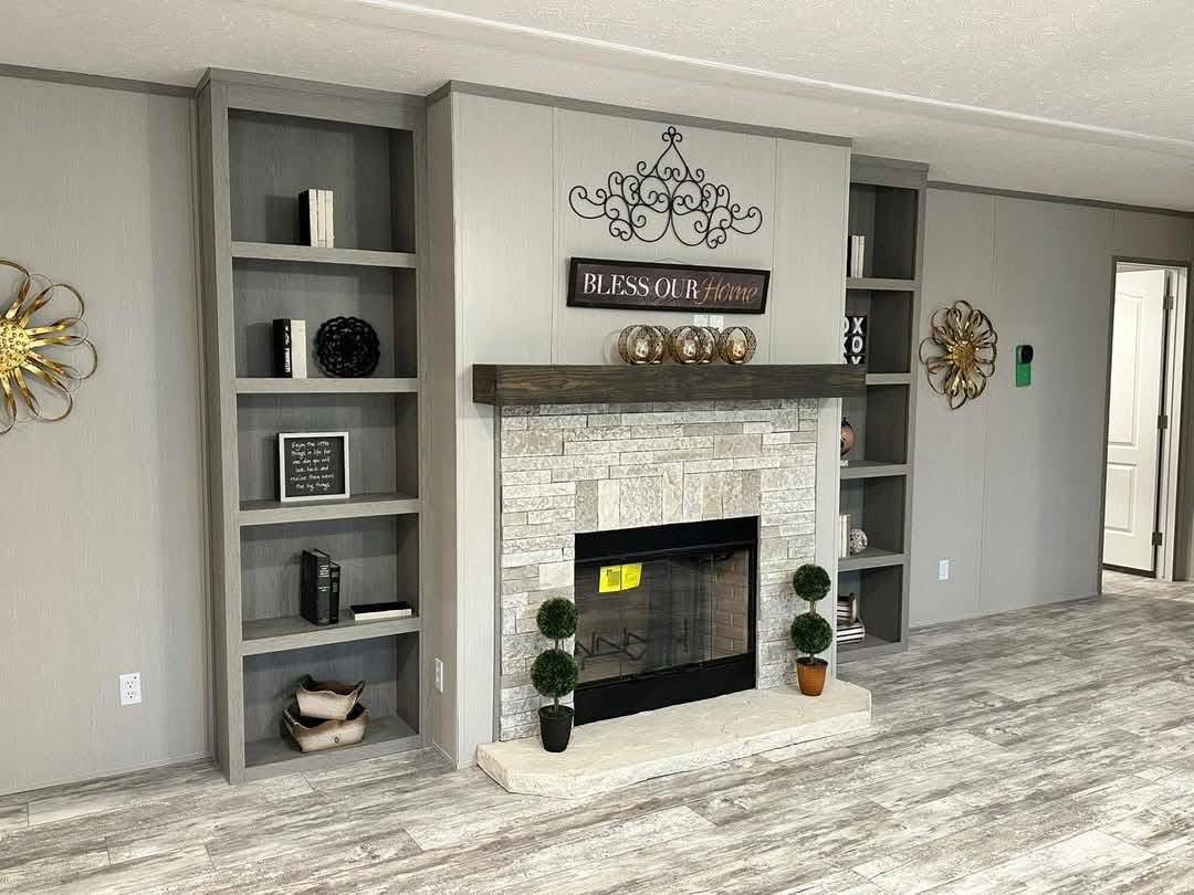 Living room with a central stone fireplace, dark wood mantel, and decorative sign reading "Bless Our Home." Flanked by gray bookshelves, cozy feel.