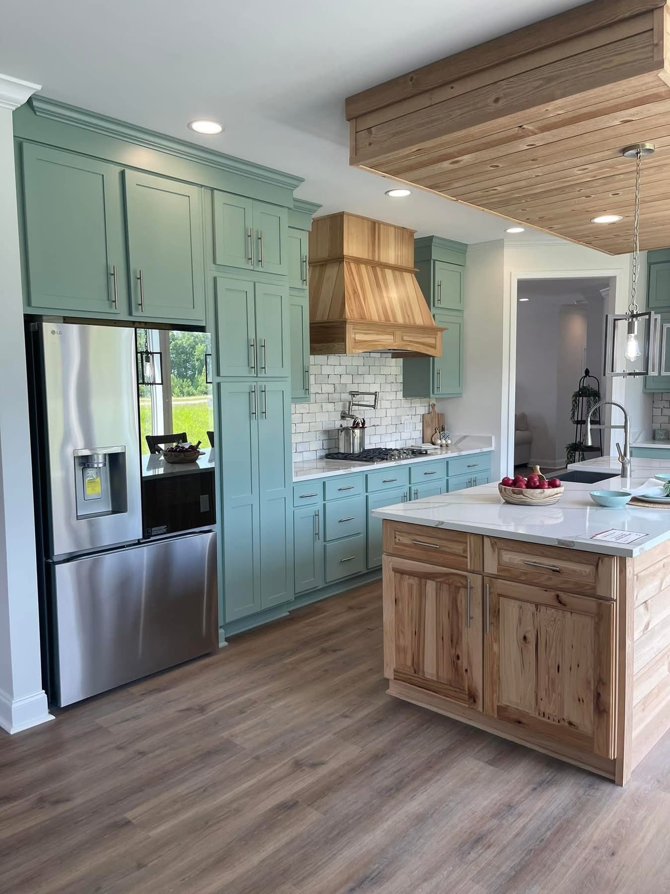 Modern kitchen with mint green cabinets, stainless steel fridge, and a wooden hood over the stove. A wood island features a white countertop with red apples.