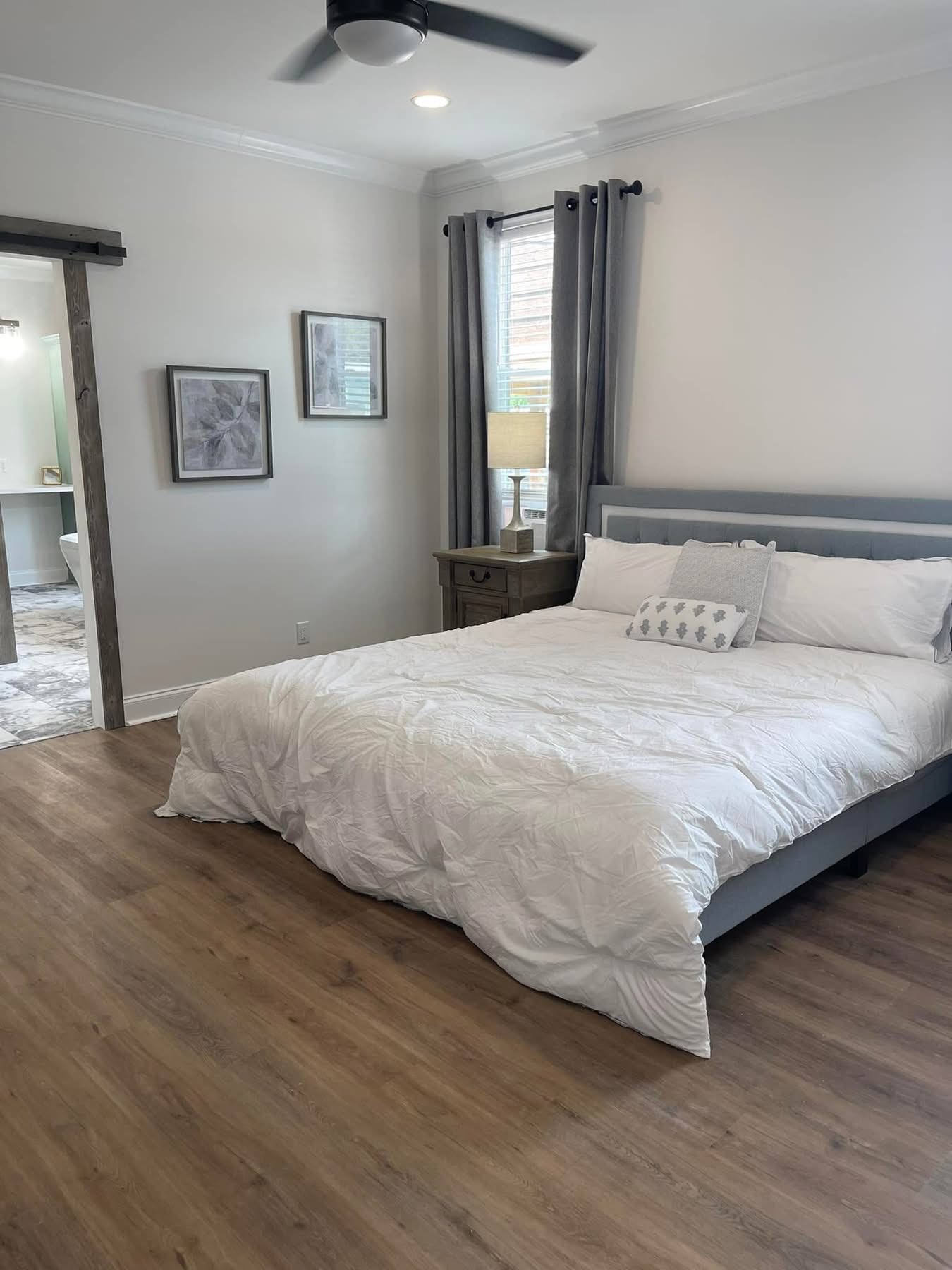 Spacious bedroom with wood flooring, a large bed with white bedding, and a pillow. Gray curtains frame a window beside a lamp on a nightstand.