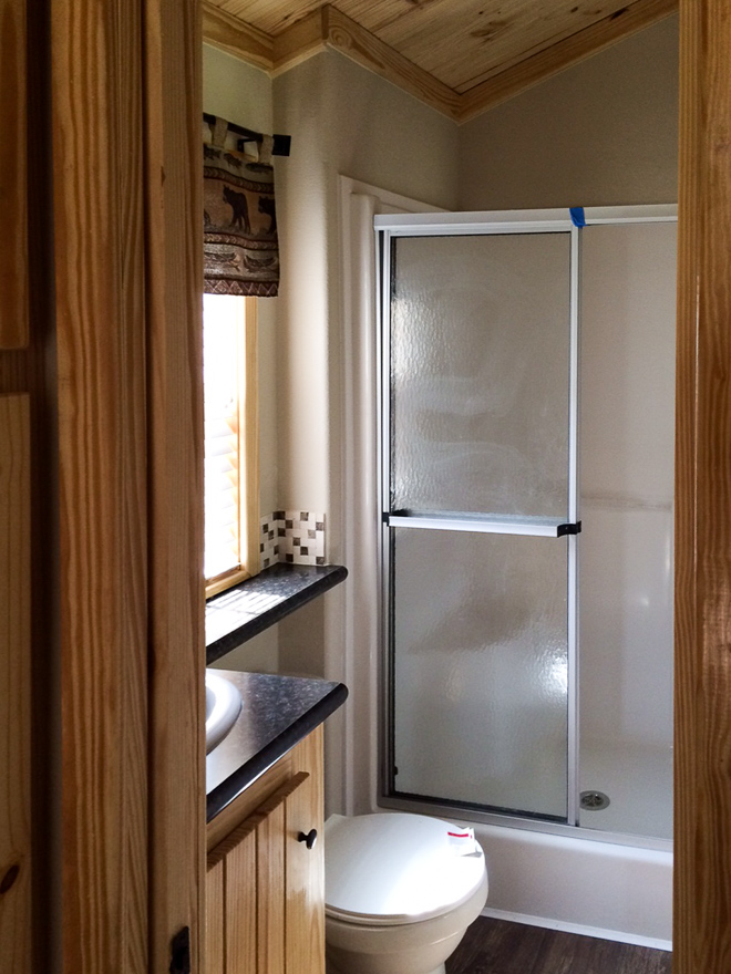 Compact rustic bathroom with wood accents, featuring a glass-door shower, white toilet, and dark countertop under a window with a wildlife-patterned valance.