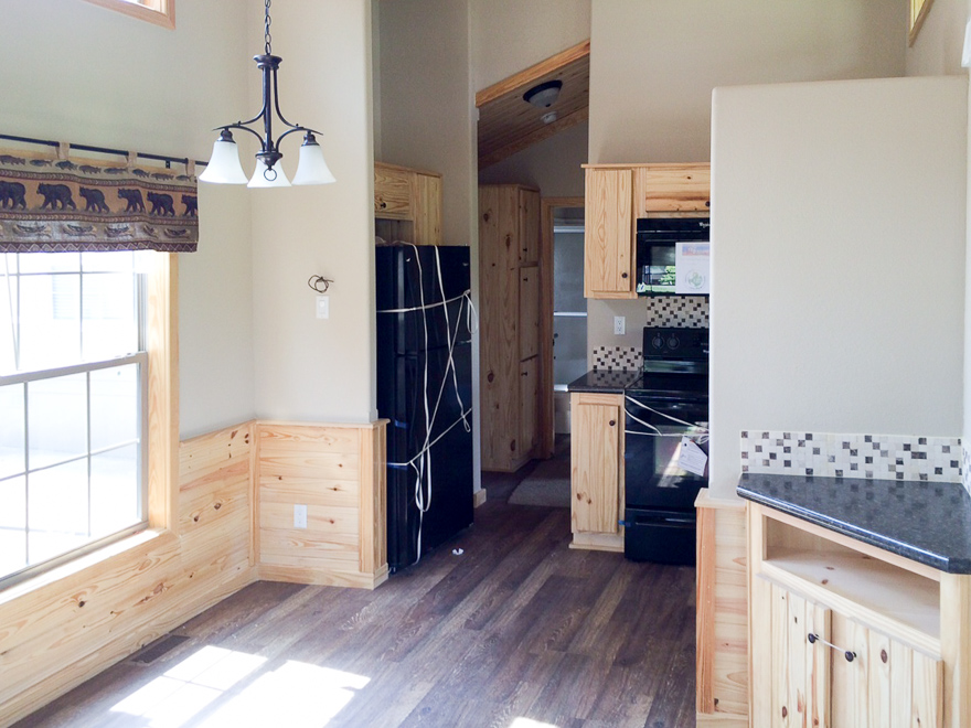 Cozy kitchen with light wood cabinetry and dark appliances. A large window with bear-patterned valance and a black chandelier add rustic charm.