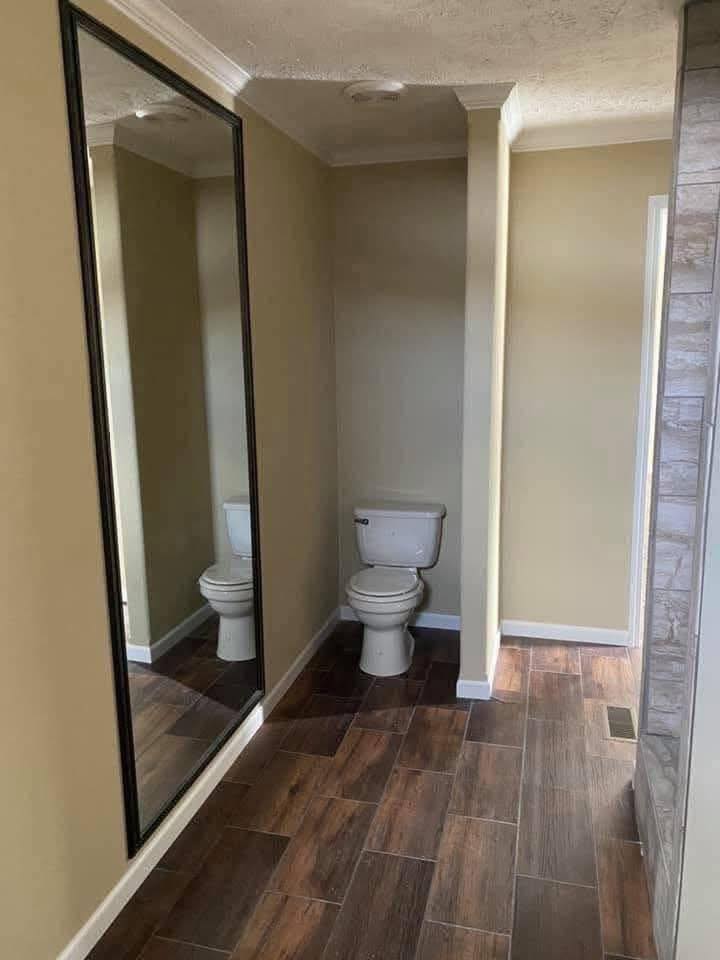 Hallway with wood-patterned tiles, featuring a large wall mirror reflecting a toilet. The scene conveys an unusual, humorous bathroom setup.