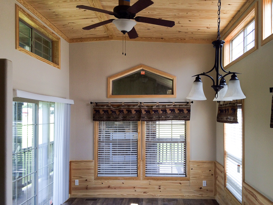 Sunlit room with wooden ceiling and floor, featuring large windows with blinds, a ceiling fan, and a chandelier. Cozy and inviting space.