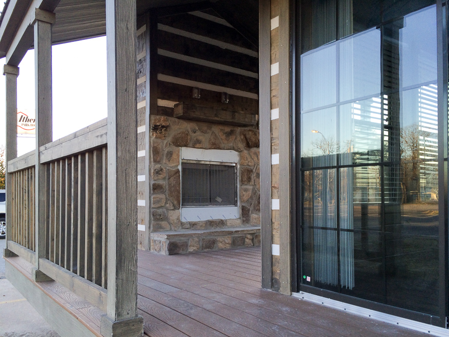 Covered porch with wooden railing and stone fireplace. Reflections visible in large glass door. Rustic, cozy ambiance with warm tones.