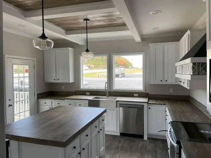 A modern kitchen with white cabinets, stainless steel appliances, and wood countertops. Two pendant lights hang above a central island. Sunlight streams through large windows, enhancing the bright, clean atmosphere.