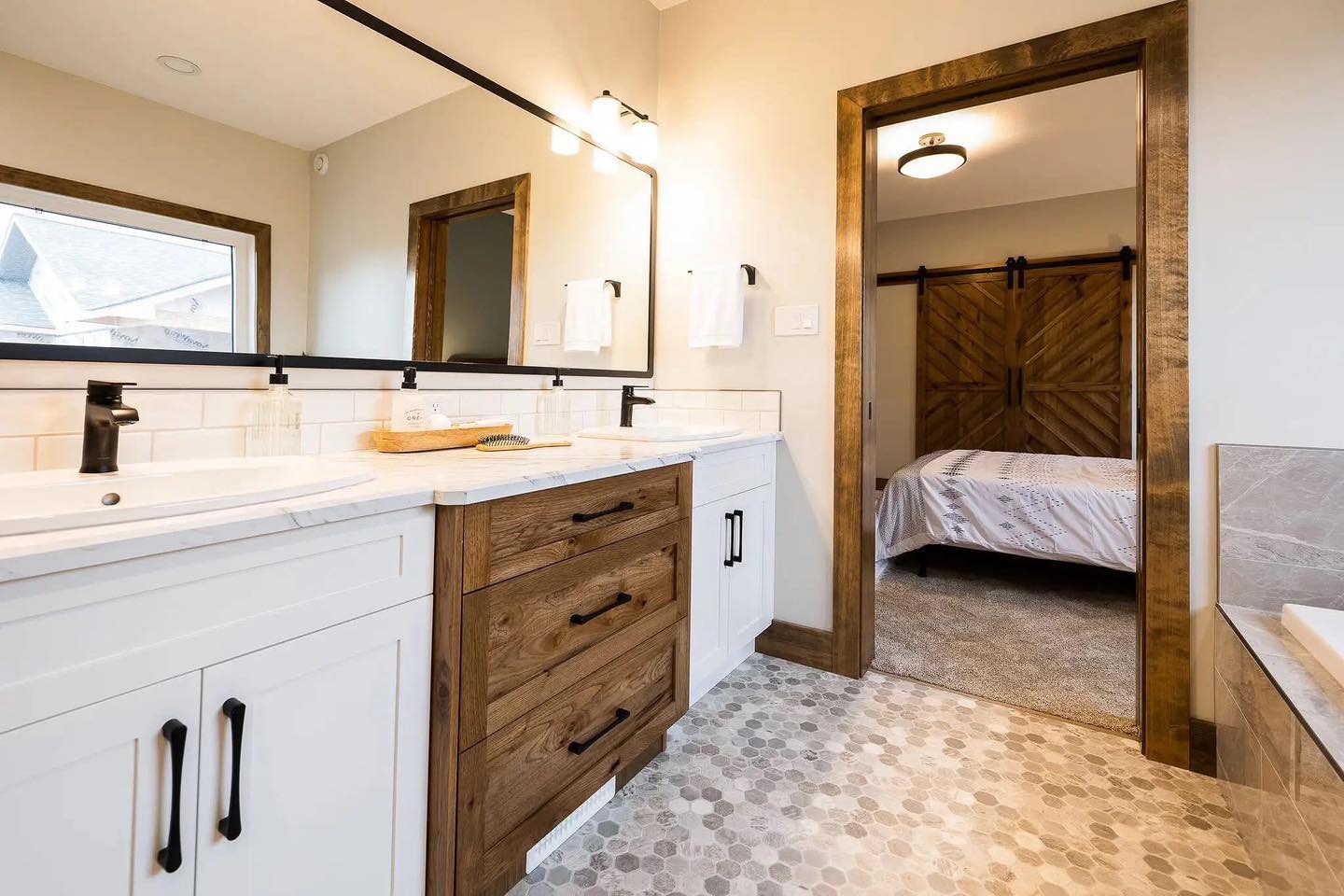 Modern bathroom with rustic charm featuring a wooden vanity with two sinks, a large mirror, and a doorway to a bedroom with a sliding barn door.
