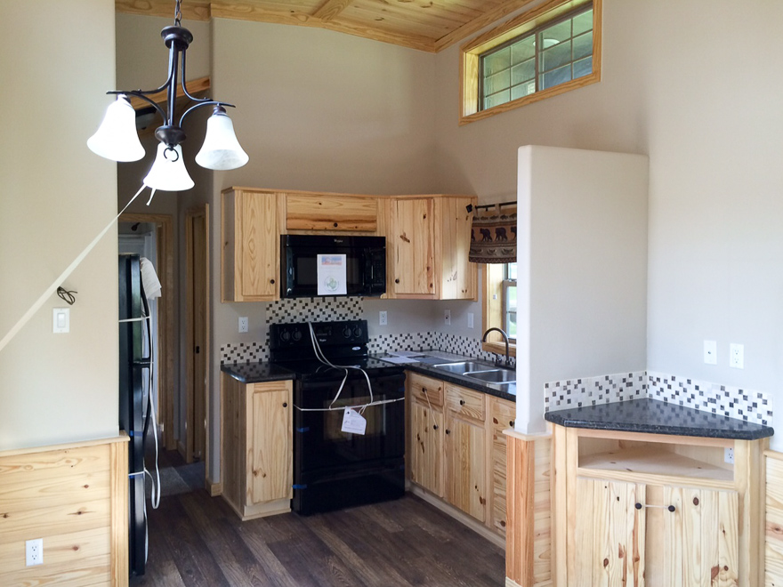 Compact kitchen with light wood cabinets, black appliances, and a mosaic tile backsplash. A modern chandelier hangs overhead, adding warmth to the space.