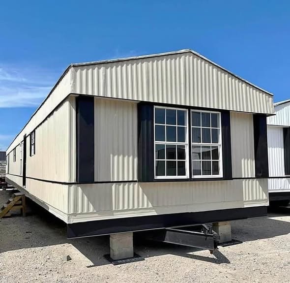 A beige mobile home with black trim sits on concrete blocks against a clear blue sky. Large windows are centered, creating a simple and functional appearance.