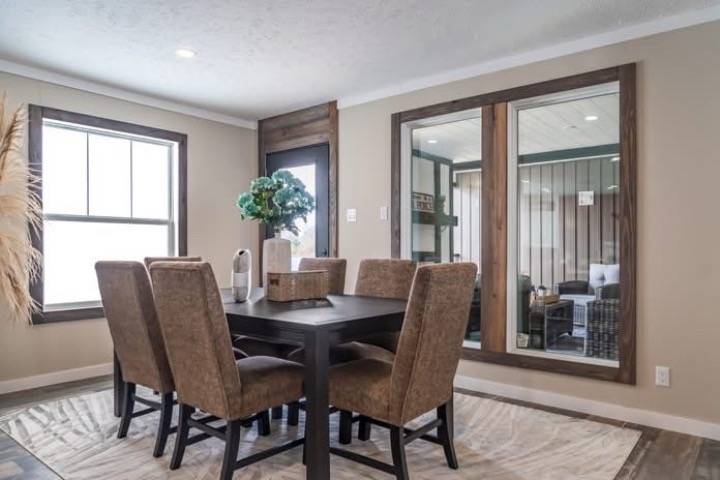 A cozy dining room features a dark wood table with six brown upholstered chairs on a light rug. Large windows and a plant add brightness and warmth.