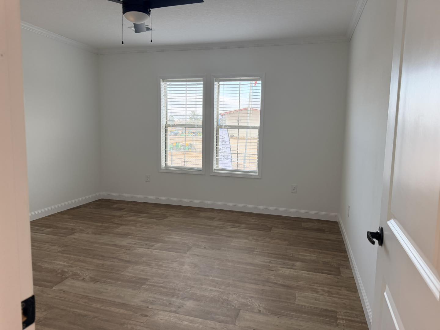 Empty room with light wood flooring, two windows with blinds, and a black ceiling fan. White walls create a bright, clean, and minimalist feel.