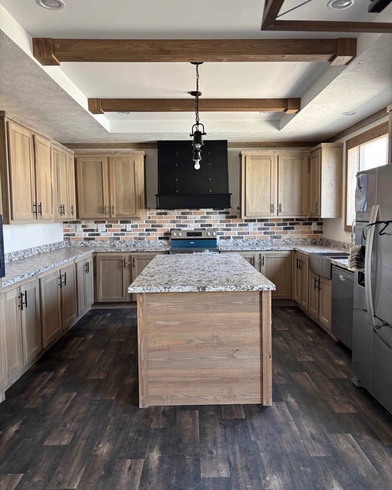 Spacious kitchen with wooden cabinets, a granite island, and a dark wood floor. Brick backsplash, black range hood, and rustic lighting create a cozy ambiance.