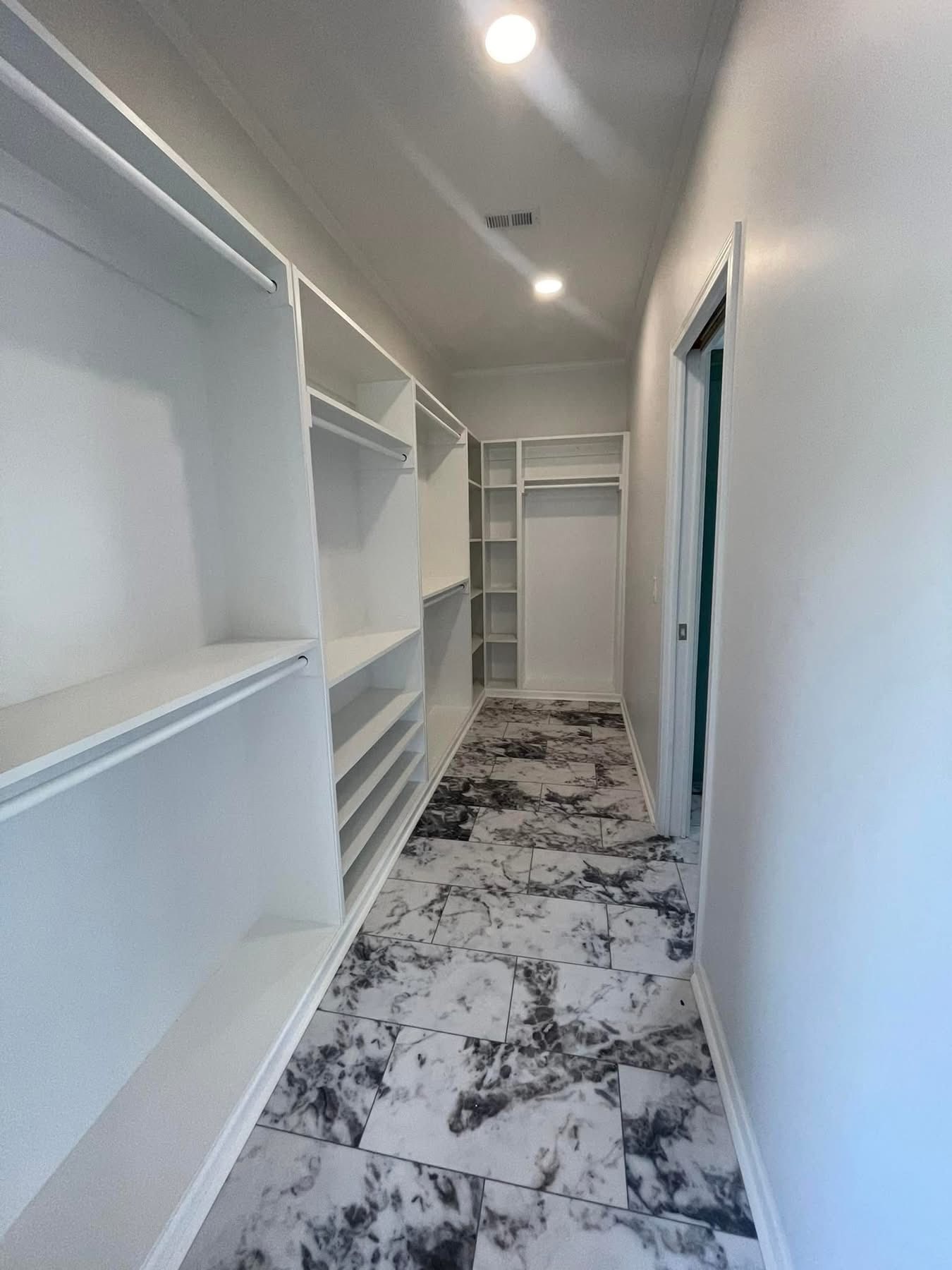 A narrow walk-in closet with white shelves on both sides and black and white marble-patterned floor tiles. Overhead lighting creates a clean, modern feel.
