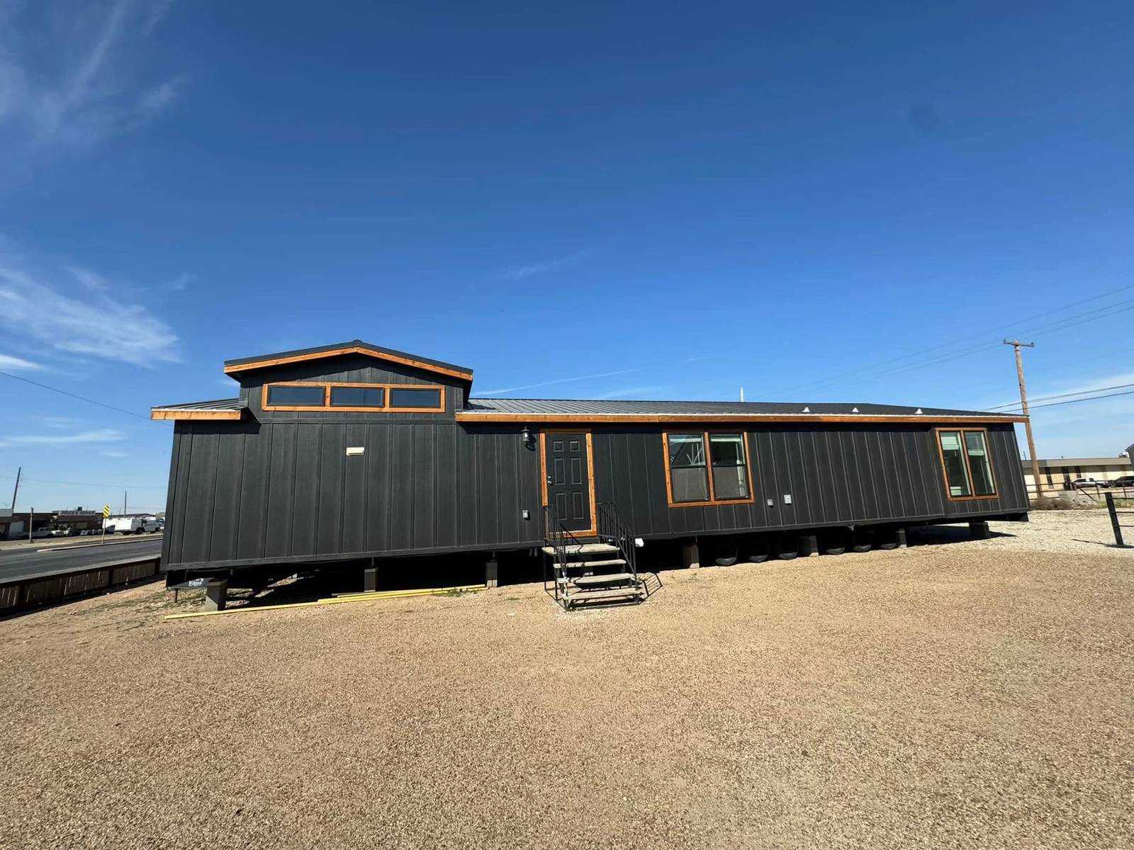 A modern, dark gray modular home stands on a gravel lot under a clear blue sky. It features large windows and a small metal stairway leading to the door.
