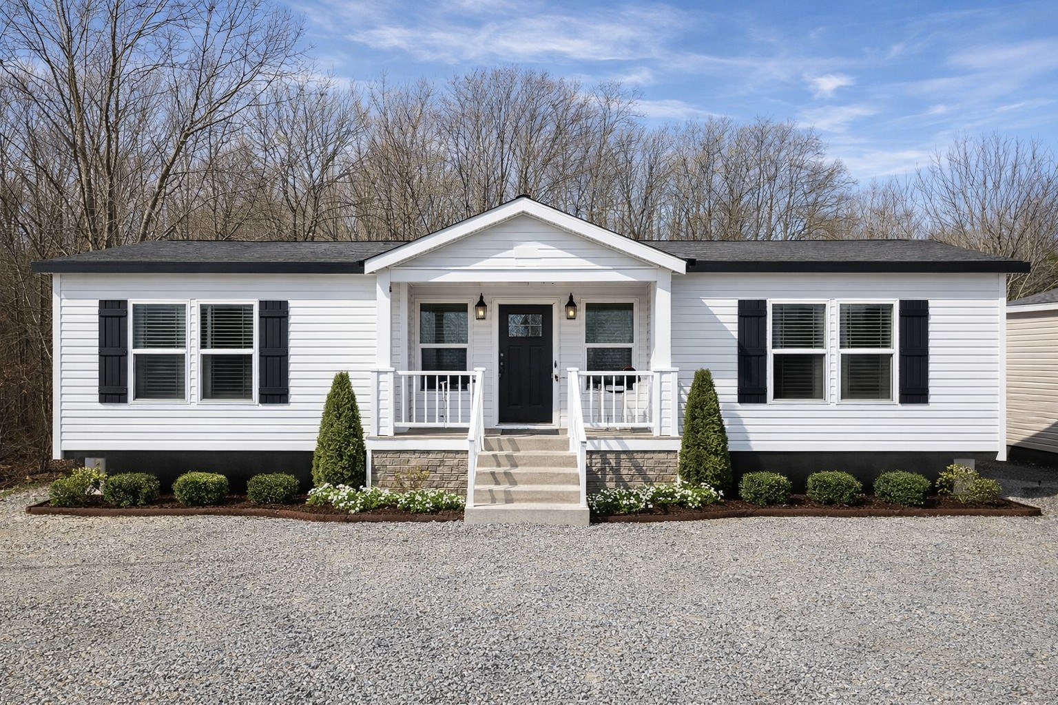 A white, single-story, ranch-style house with black shutters and a black door, set amidst bare trees under a blue sky. Neatly landscaped with shrubs.