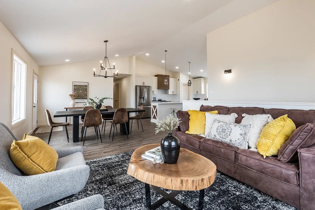 Spacious living room with neutral tones, featuring a brown sofa with yellow cushions, a rustic wooden coffee table, and a chandelier over a dining table.
