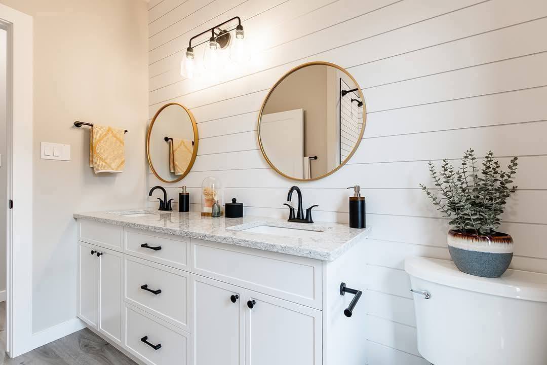 Modern bathroom with white shiplap walls, dual round mirrors above a marble countertop, black fixtures, a potted plant on the toilet, and soft lighting.