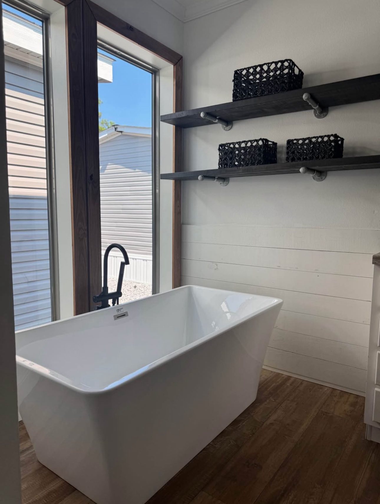 A modern bathroom features a white rectangular bathtub beside large windows with wooden frames. Two shelves hold black wicker baskets. The scene conveys tranquility.