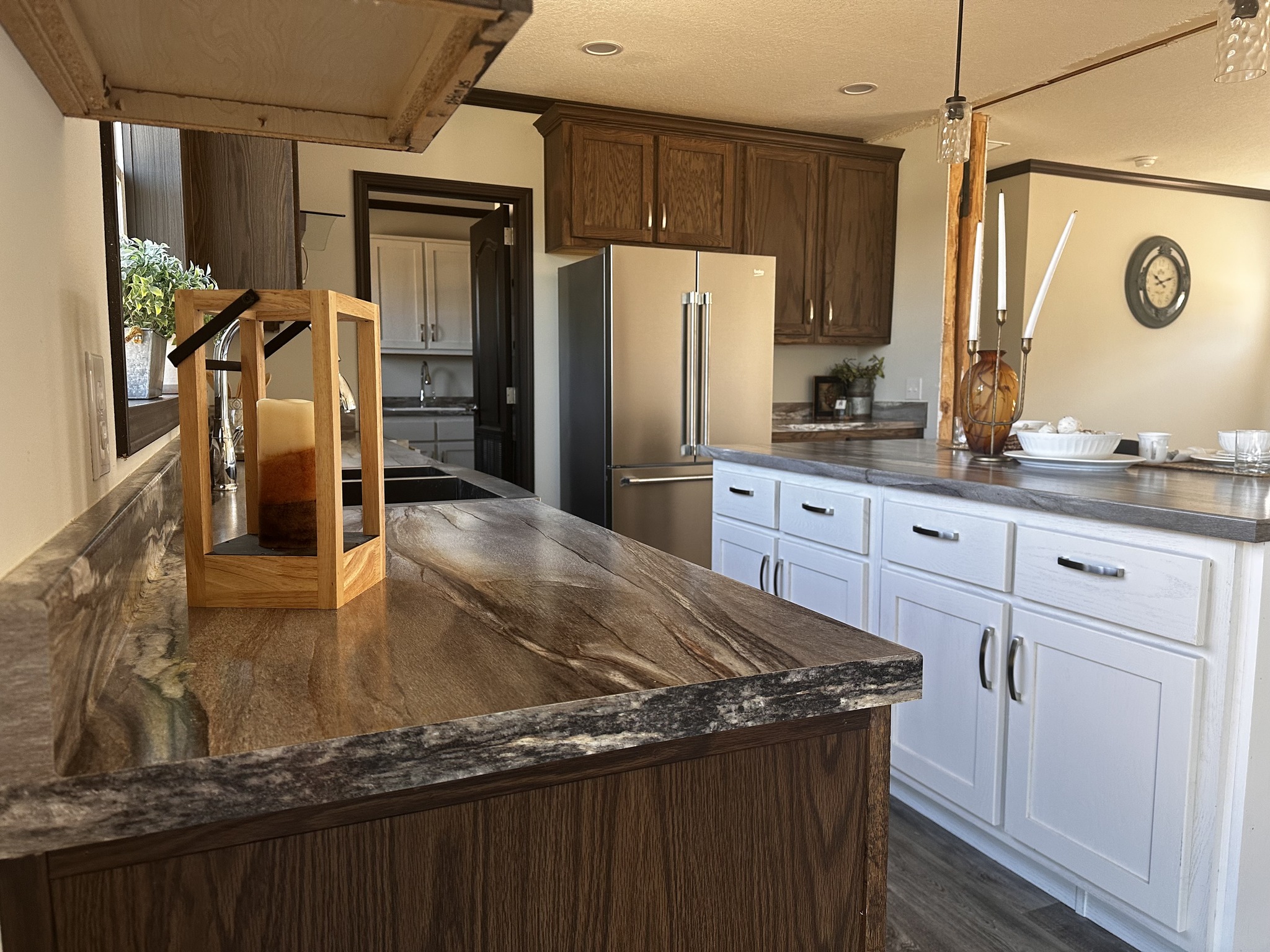 Modern kitchen with a warm decor. Features marble countertops, a stainless steel fridge, and wooden cabinets. A candle and tableware add cozy accents.