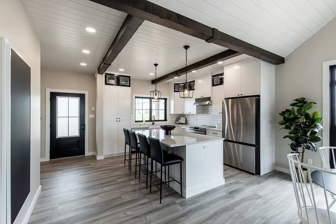 Modern kitchen with a white island, black barstools, and pendant lights. Dark wooden beams contrast against a white ceiling, creating a cozy, stylish vibe.