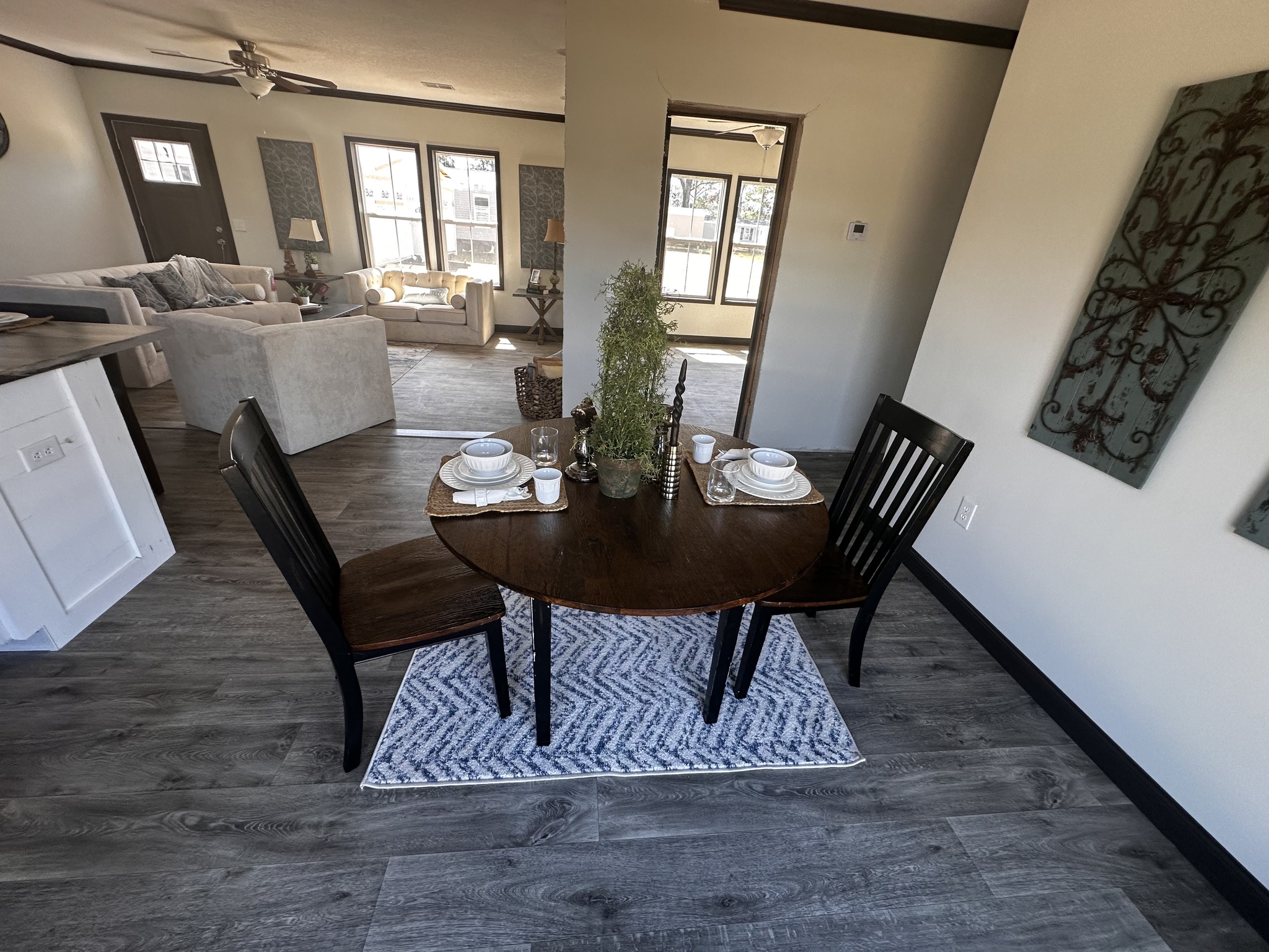 Cozy dining area with a dark wooden table set for two on a patterned rug. Bright living room with cushioned sofas and large windows in the background.