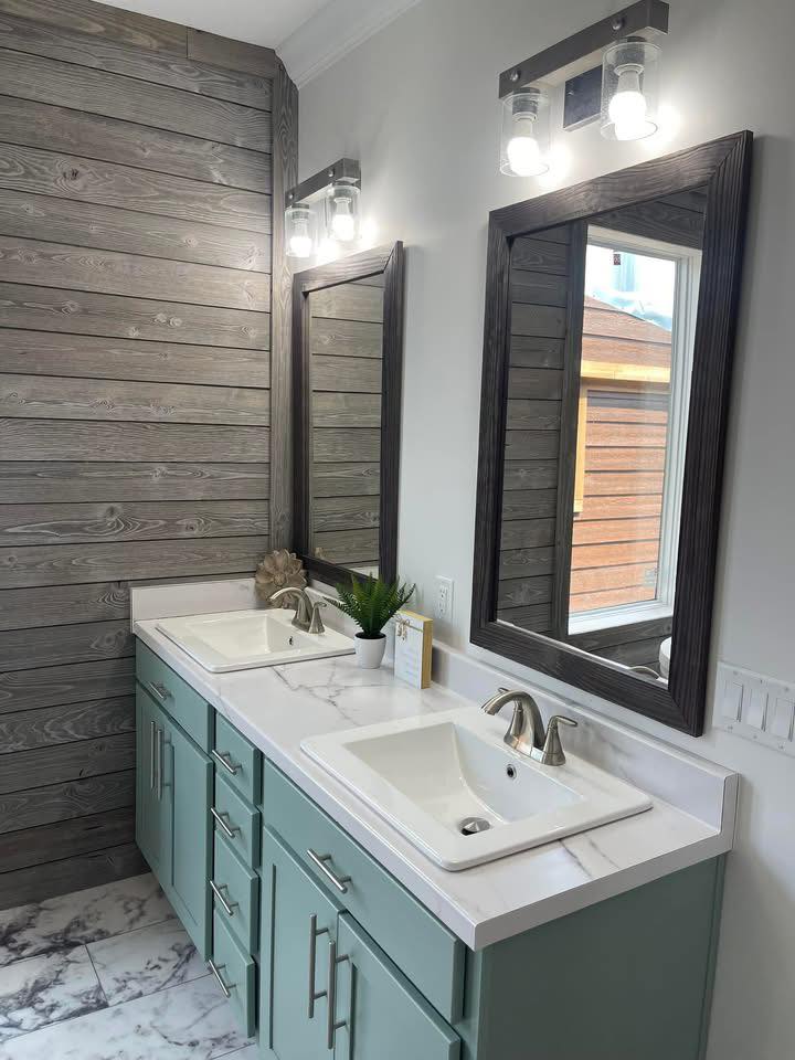 Modern bathroom with dual white sinks set in a marble countertop on teal cabinets. Two large mirrors and industrial lights enhance the rustic wood wall.