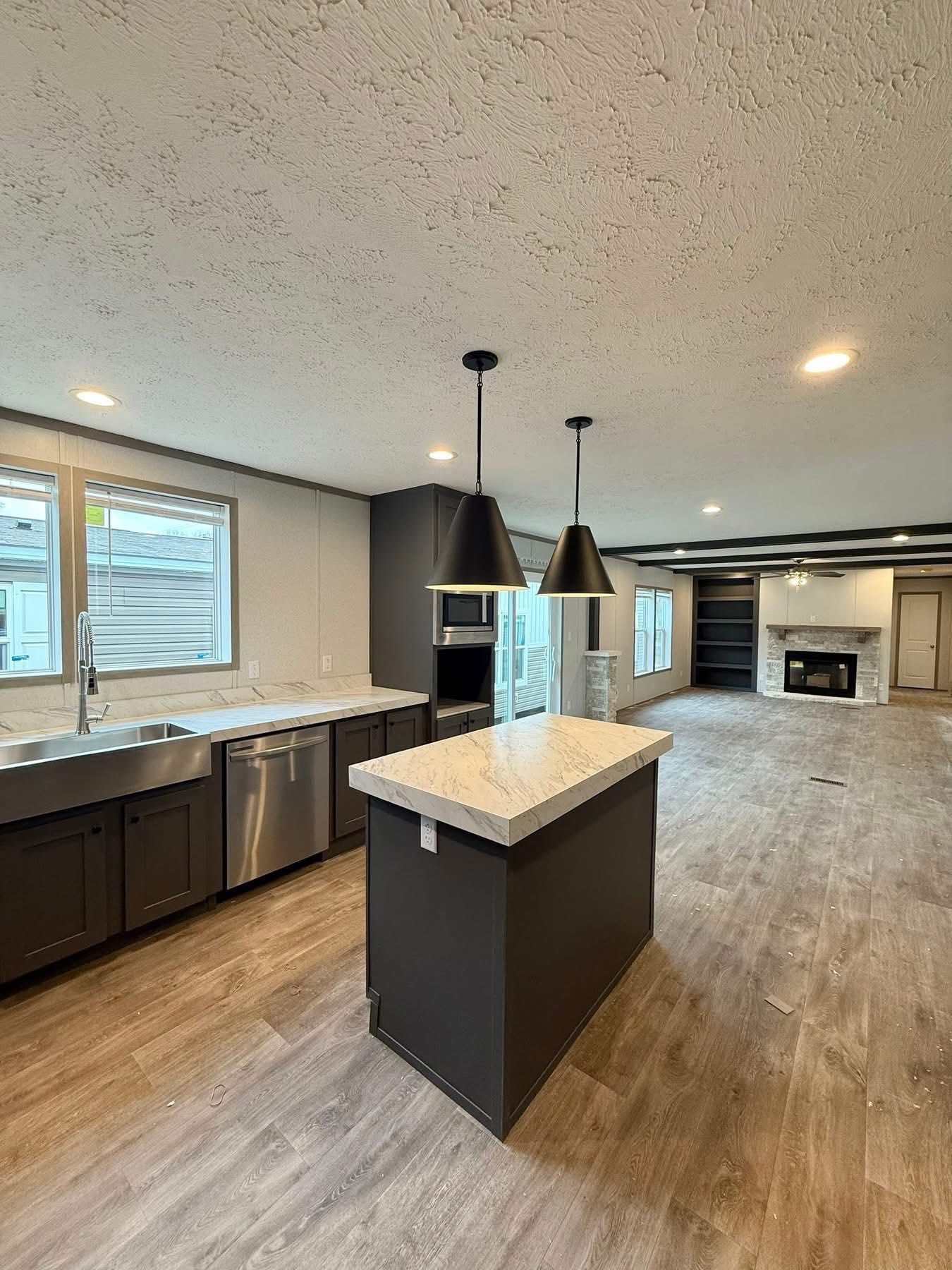 Modern open-plan kitchen and living area with a large island, marble countertops, dark cabinets, wood floors, hanging pendant lights, and a cozy fireplace.